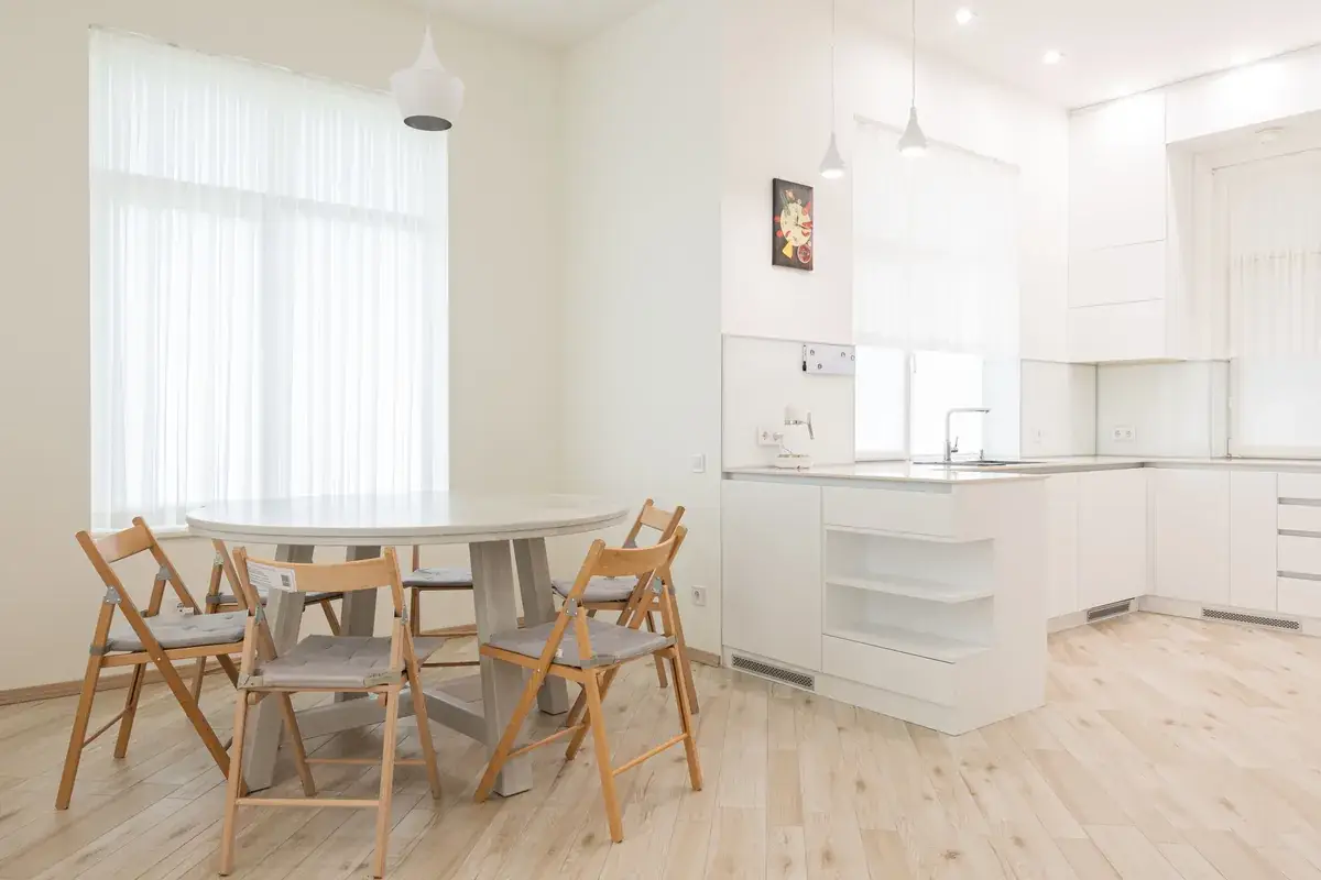 Round white dining table with four wooden folding chairs in a white kitchen with sheer curtains