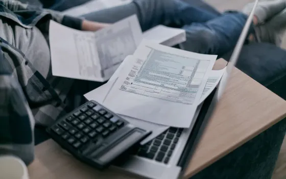 Person in a checked shirt sitting cross-legged on a sofa with a calculator, a laptop and a sheaf of printed forms on a wooden tray