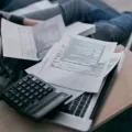 Person in a checked shirt sitting cross-legged on a sofa with a calculator, a laptop and a sheaf of printed forms on a wooden tray