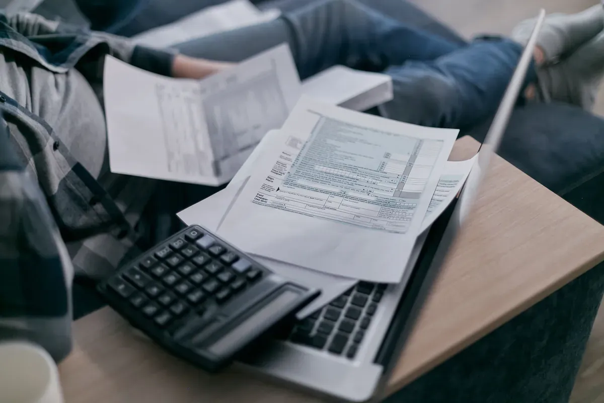 Person in a checked shirt sitting cross-legged on a sofa with a calculator, a laptop and a sheaf of printed forms on a wooden tray