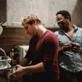 Man in a maroon top rinsing a steel pan at a kitchen sink while a friend in a denim shirt holds a mug and laughs