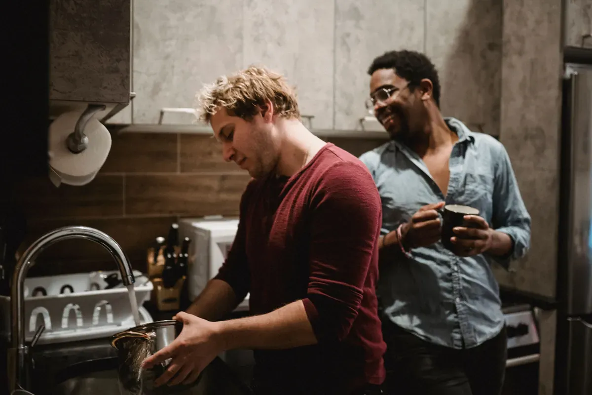 Man in a maroon top rinsing a steel pan at a kitchen sink while a friend in a denim shirt holds a mug and laughs