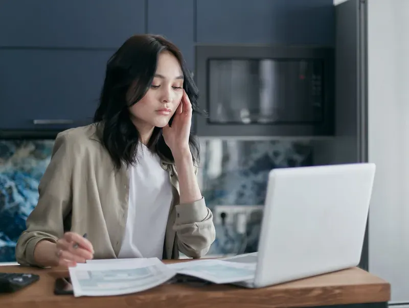 Dark-haired woman with a pen in hand, head resting against her fingers, studying open papers beside a laptop