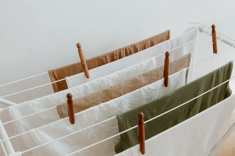White folding clothes airer with tea towels and a green cloth pegged to its rails with wooden pegs, against a white wall