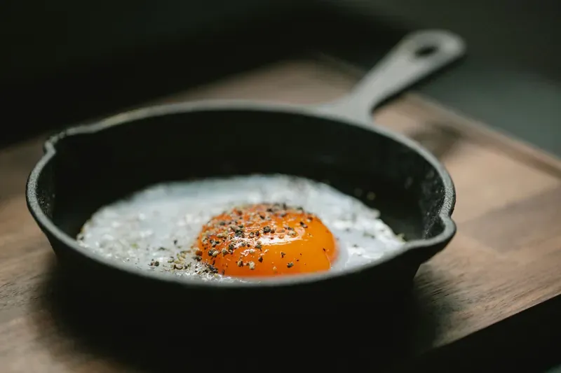 Cast-iron frying pan on a wooden board with a single fried egg, its orange yolk cracked with black pepper