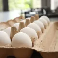 Open cardboard egg box on a worktop with white eggs and a small feather, blurred garden light behind