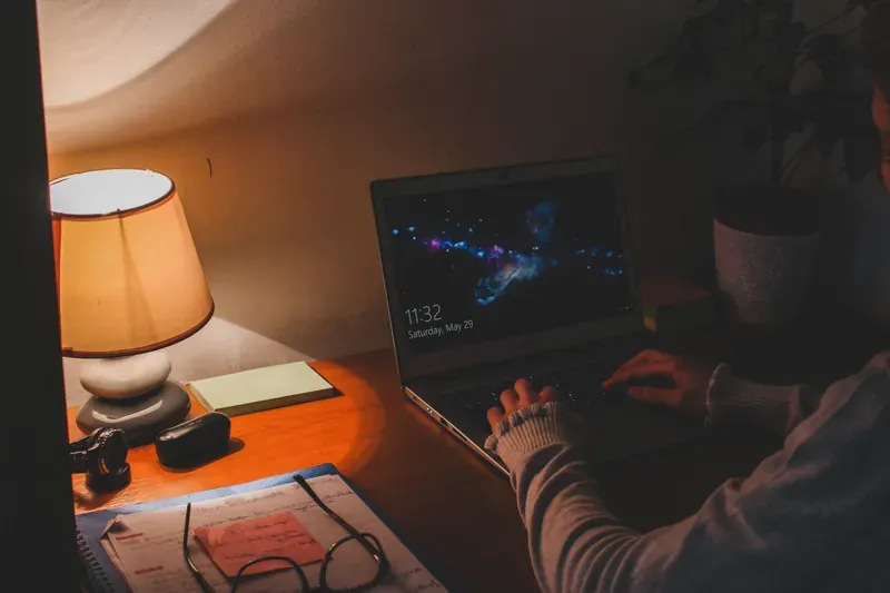 Laptop on a wooden desk at night showing 11:32 on its lock screen, lit by a small table lamp beside a notebook and glasses