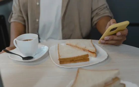 Person in a beige shirt holding a yellow-cased phone beside a cup of tea and a plate of cut sandwiches