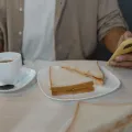 Person in a beige shirt holding a yellow-cased phone beside a cup of tea and a plate of cut sandwiches