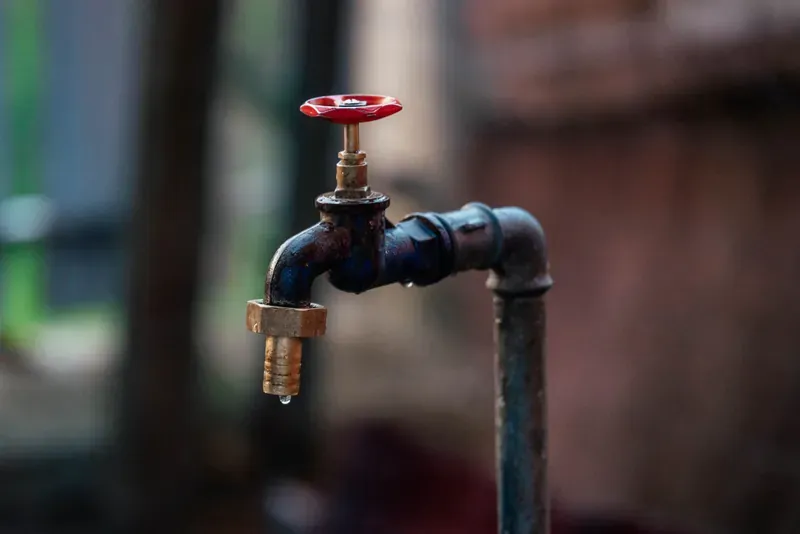 Outdoor tap with a red wheel valve and a single water droplet hanging from the spout