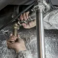 Two gloved hands using a wrench to tighten a large brass pipe fitting against foil-wrapped insulation