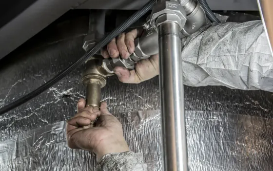 Two gloved hands using a wrench to tighten a large brass pipe fitting against foil-wrapped insulation
