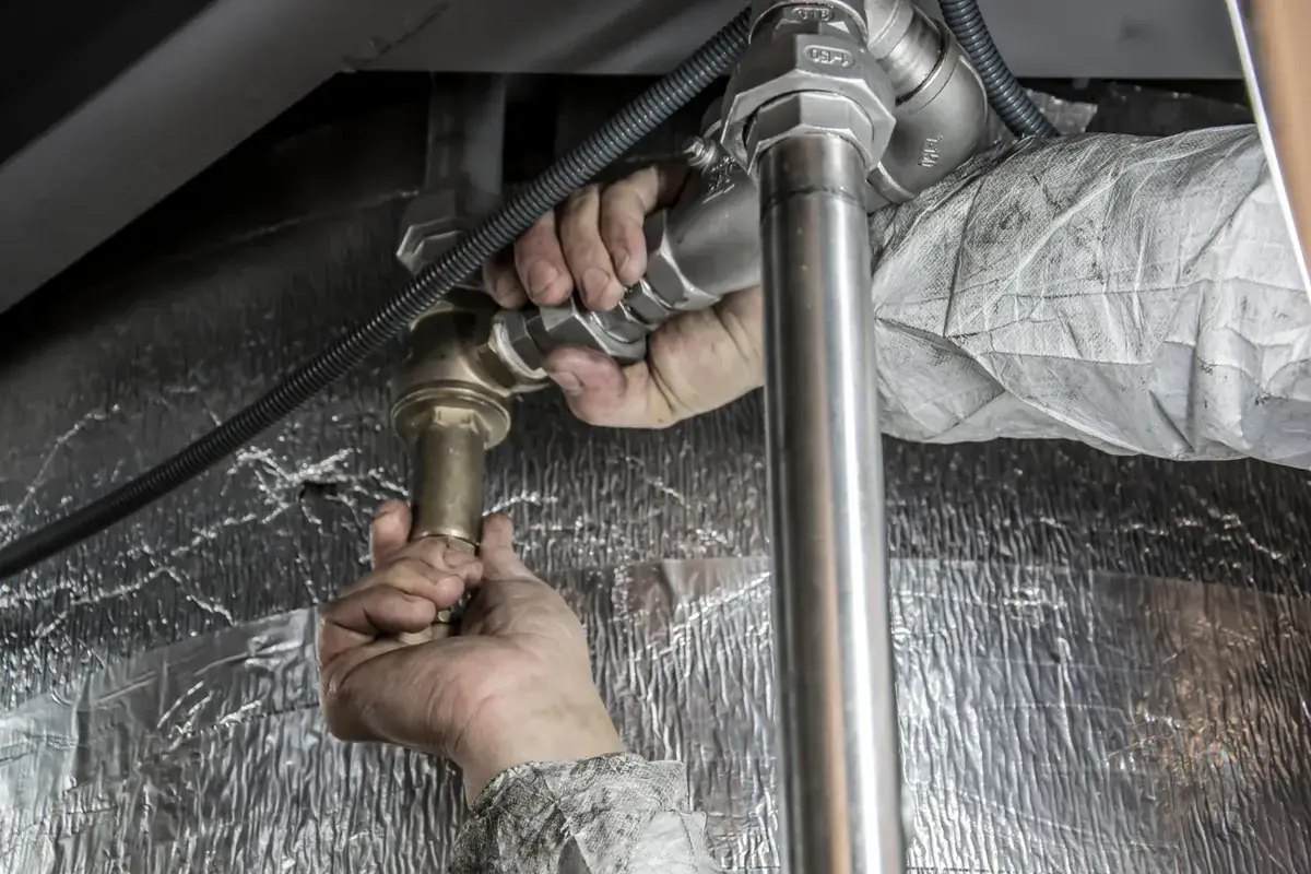 Two gloved hands using a wrench to tighten a large brass pipe fitting against foil-wrapped insulation