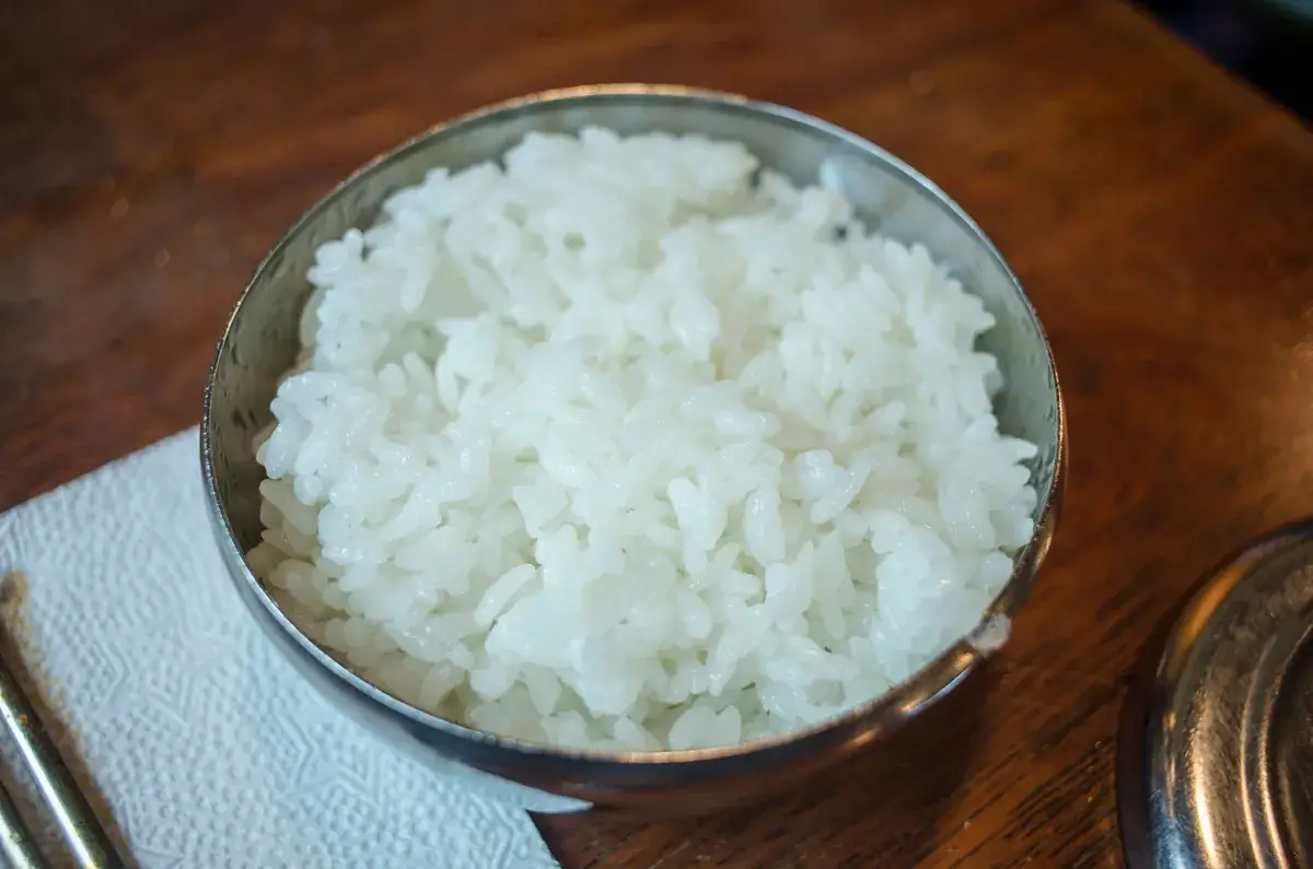 Steel bowl of plain white rice on a dark wooden table beside a folded paper napkin
