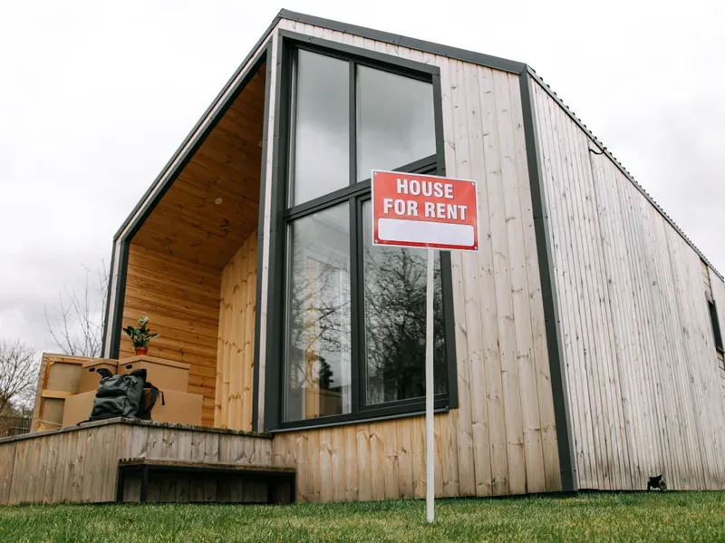 Modern timber-clad house with a red HOUSE FOR RENT sign on a post, moving boxes stacked on the porch