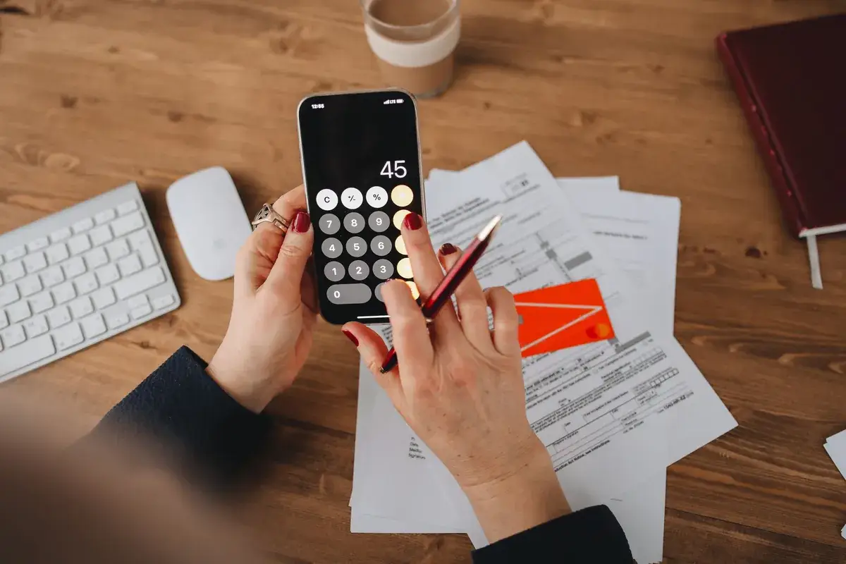 Hands holding a phone calculator showing the number 45, over bills and a bank card on a wooden desk