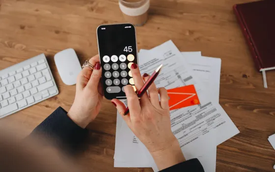 Hands holding a phone calculator showing the number 45, over bills and a bank card on a wooden desk
