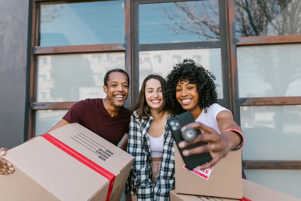 Three friends smiling for a phone selfie while holding moving boxes outside a building