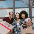 Three friends smiling for a phone selfie while holding moving boxes outside a building