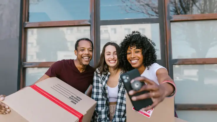 Three friends smiling for a phone selfie while holding moving boxes outside a building