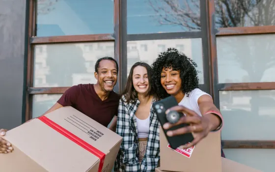Three friends smiling for a phone selfie while holding moving boxes outside a building