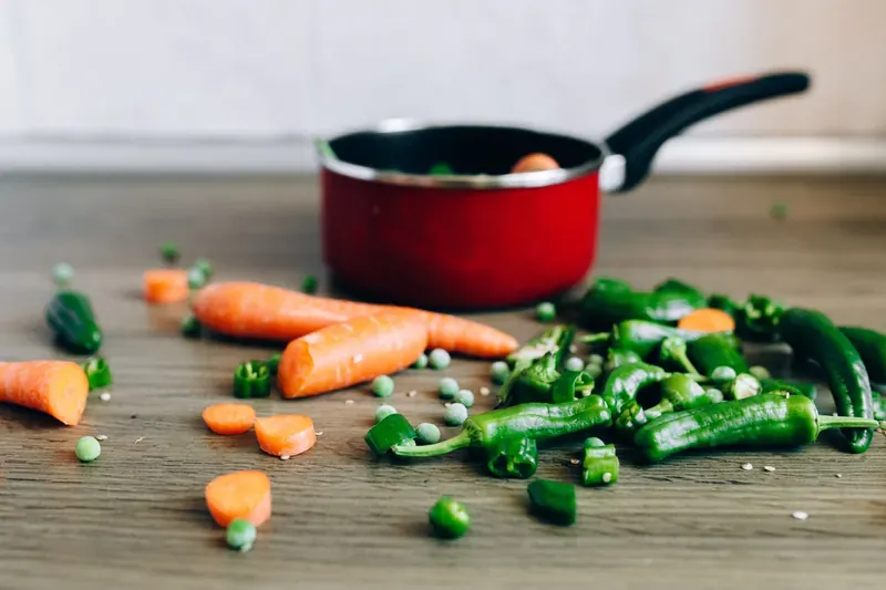 Carrots, chillies and loose peas scattered on a wooden counter beside a red saucepan