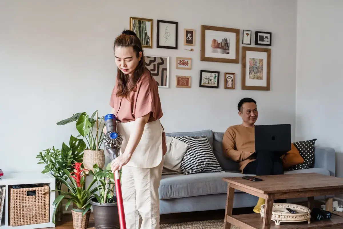Woman vacuuming a living room with houseplants while a man sits on a sofa using a laptop
