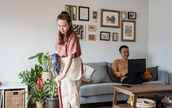 Woman vacuuming a living room with houseplants while a man sits on a sofa using a laptop
