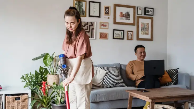 Woman vacuuming a living room with houseplants while a man sits on a sofa using a laptop