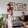 Woman vacuuming a living room with houseplants while a man sits on a sofa using a laptop