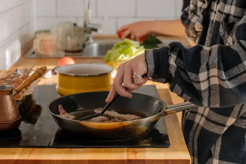 Person cooking bacon in a frying pan on a hob, with eggs and vegetables on the worktop nearby