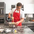 Teenager in a red apron whisking ingredients in a steel mixing bowl beside eggs and butter on a kitchen island