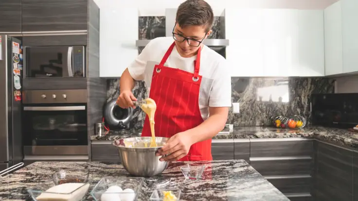 Teenager in a red apron whisking ingredients in a steel mixing bowl beside eggs and butter on a kitchen island