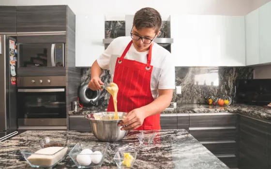 Teenager in a red apron whisking ingredients in a steel mixing bowl beside eggs and butter on a kitchen island