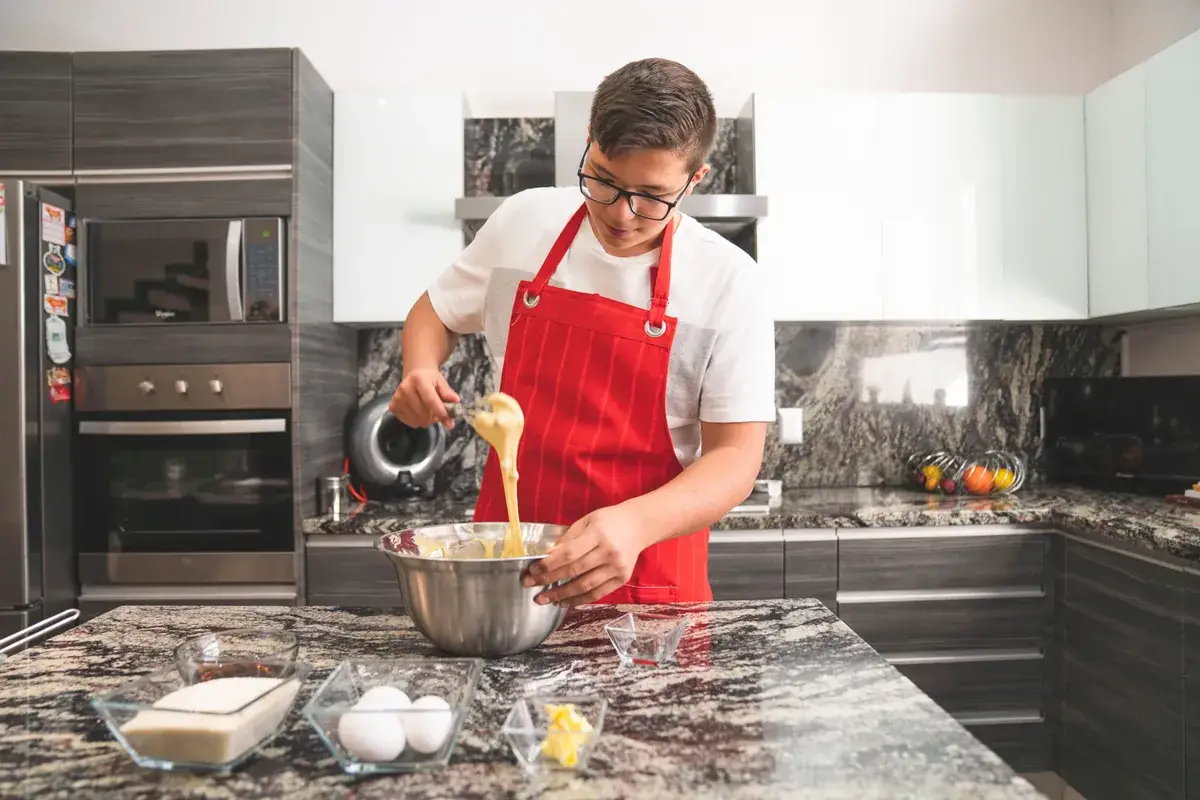 Teenager in a red apron whisking ingredients in a steel mixing bowl beside eggs and butter on a kitchen island